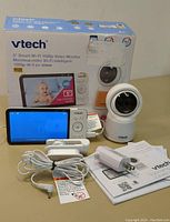 Wide view showing VTech branded 5" Smart Wi-Fi 1080p video baby monitor box, monitor screen, camera, power adapters, and manuals in front of box.