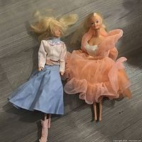 Two vintage Barbie dolls laid on a floor, one in pink and blue outfit and the other in peach-pink dress.
