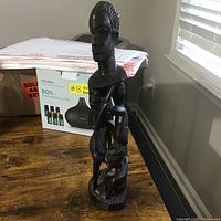 Tall dark wood carving showing elongated standing human figure on top and two smaller figures near the base, on a wooden table.