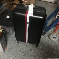 Black hard body suitcase with a red and white vertical stripe on the front, standing upright on four wheels on a speckled floor, with a shipping tag attached.