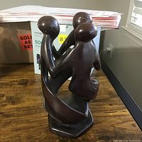 Carved wooden statue showing abstract figures from one side, placed on wood floor with cardboard and boxes in background