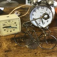 Front view of two clocks - Sharp twin bell alarm clock and cream-colored electric alarm clock, with wire bicycle figurine in front.
