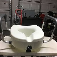 Front view of the white plastic Fodac toilet seat with black adjustment knob and silver side handles with black grips.