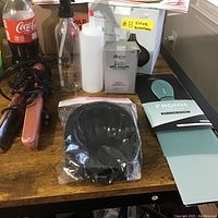 Lot of hair care products and tools on wood surface including flat iron, spray bottles, black manicure bowl, and boxed hair end wraps.