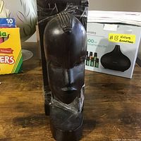 Front view of one hand carved African wooden bookend showing facial features and hair detail, placed on table with background items.