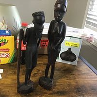 Front view of the African man and woman wooden carvings on a table with household items in the background.