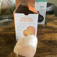 Heart-shaped Himalayan LED salt lamp wrapped in plastic, showing hand-carved light pinkish salt on a round wooden base in front of its original box explaining features.