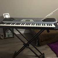 Front angled view of Casio CTK-491 keyboard on black X-style stand placed in room corner on hardwood floor with visible small crack on bottom of keyboard.