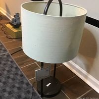 Brown metal table lamp with beige fabric shade, tag attached on base, shown from two angles on floor near wall.