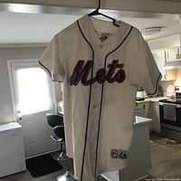 Front view of white Mets baseball jersey hanging on hanger showing front team name and button closure with blue piping.