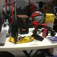Overview of miscellaneous items on white table including vacuum, speakers, rat trap, mirror, towing strap, and small boxes