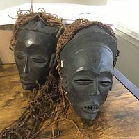 Two hand-carved African wooden masks displayed on a wooden table, showing full front and side angles with hair details.