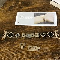 Gold-tone watch band with black clover-shaped insets and rhinestones laid on wooden table along with two extra metal link pieces and an installation instruction sheet showing watch band worn on wrist.