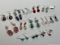 Full view of all 14 pairs of assorted semi-precious stone earrings arranged on white background, showing variety of colors and styles.