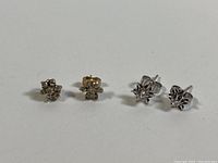 Two pairs of small stud earrings displayed front-facing: yellow gold cluster style with six melee diamonds per earring and white gold elevated single diamond studs.