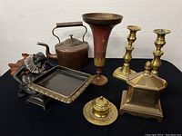 Seven brass and cast iron items including monkey butler tray, lidded tobacco box, trumpet vase, candlesticks, inkwell, kettle, and brass container arranged on black fabric