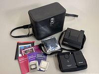 Full lot overview showing Minolta Instant PRO camera, Polaroid Spectra System remote control, manuals, lens filters, sealed film pack, and black leather carrying case on table.