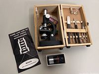 Tasco microscope inside the open wooden box with tools in compartments and original manual beside it. Celestron lens in plastic case below.