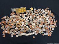 Wide view showing a large assortment of scattered coins of various origins on a black surface, including a boxed 1988 Korea Olympics commemorative coin set with gold and silver coins and a coin bracelet.