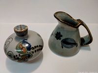 Two ceramic pieces side by side: a small water pitcher and an oil dispenser container, both with floral and leaf painted decoration.