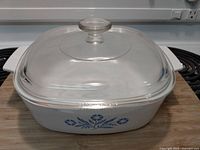 Square CorningWare Blue Cornflower casserole dish with clear Pyrex glass lid on a wooden cutting board, shown from front angle.