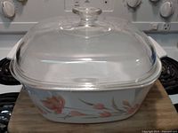 Square Corning Ware casserole dish with floral design and Pyrex glass lid sitting on stove.