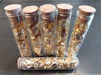 Photo showing six small clear glass vials filled with gold leaf flakes and sealed with cork stoppers, arranged upright with one vial horizontal in front.