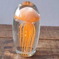 Full view of clear glass paperweight with internal orange jellyfish design on wooden surface.