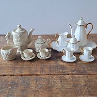 Two sets of miniature porcelain tea sets on wooden table; one set white porcelain with gold trim, one set off-white with floral embossing.