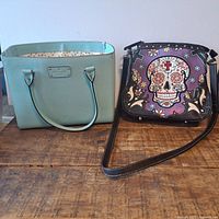 Two Kate Spade purses on wooden surface: light blue tote and purple skull pattern crossbody