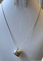 Image showing shiny sterling silver heart-shaped pendant on a thin 16-inch silver chain displayed on a white bust stand.