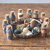 All labeled glass bottles filled with different polished gemstone chips arranged in a semi-circle around a metal dish containing loose polished stones.