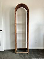 Front view of the tall arched wicker rattan etagere with four clear Lucite shelves against a white wall.