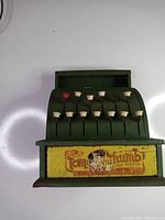 Front view of the green metal Tom Thumb toy cash register showing two rows of cream colored keys on metal rods with one red key, and the original yellow Tom Thumb label with cartoon character and text.