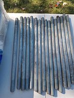 Close-up photo of 15 galvanized steel anchor rods laid in a row on a white table outdoors, showing threaded ends.