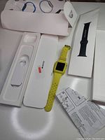 Apple Watch with yellow sport band shown next to original white Apple Watch packaging and extra black sport band box, instruction manual also visible.