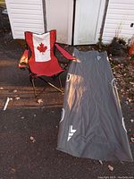 Photo showing red folding camping chair with Canadian flag design alongside gray Backwoods camping cot on pavement with leaves.