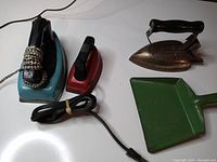 Four vintage children's toy cleaning items: a blue electric toy iron with black and white cord, a smaller red toy iron, a metal non-electric iron with black handle, and a green metal toy dustpan.
