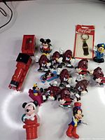 Photo showing a variety of small vintage collectible toys including Thomas the Train red engine, multiple California Raisins figures, Mickey Mouse figurines, and a Coca-Cola keychain still in packaging.