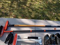 Photo showing 8 Ender Duke curling broom with orange brush head, BalancePlus Lightspeed brush broom with orange and blue handle, white Edge Glider ice scraper, curling glove, and two pairs of curling shoes (men's and women's sizes) outdoors on a table.