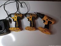Three DeWalt cordless drills with visible wear and dirt, placed side by side on a white surface alongside a DeWalt battery tester.