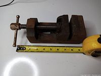 Small Eron metal bench vise alongside a measuring tape showing length approximately 7.5 inches. Surface rust visible on the screw handle and jaws.
