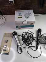 Photo showing three microphones, one with 'ON AIR' label and the WiFi security camera in box, and a wireless Bluetooth audio receiver in packaging