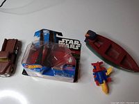 Photo showing Star Wars Hot Wheels shuttle toy in original packaging, small brown toy truck, souvenir boat, and small Burger King Transformer toy together on white background.