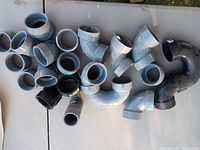 Top-down view of varied plumbing pipe fittings laid out on a table, includes elbows, tees, couplings, primarily PVC and some black fittings.