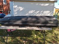 Front and side view of the large black Thule Frontier car roof carrier sitting on a folding table outside on grass. Shows overall shape, Thule logo on side, and locking mechanism on the bottom edge.