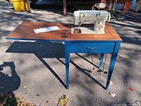 Overall view of the sewing machine built into a blue table with an extendable wooden surface and a drawer under the machine.