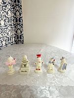 Full set of 5 Department 56 figurines displayed on white lace tablecloth in front of window with patterned curtains.