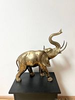 Full view of the brass elephant sculpture standing on a black platform against a white wall.