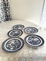Five Churchill Blue Willow dinner plates arranged on a white patterned tablecloth, showing their blue oriental-style transferware design.